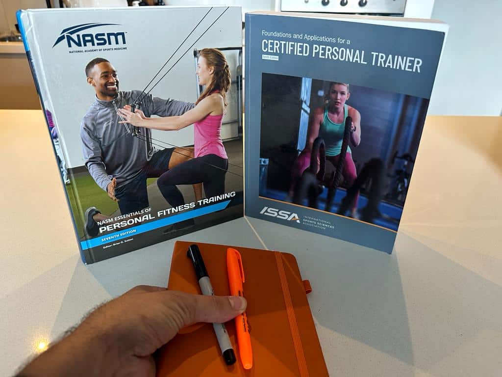 NASM cpt and ISSA cpt textbooks standing up and showing their covers while a learner holds a journal with a sharpie and highlighter