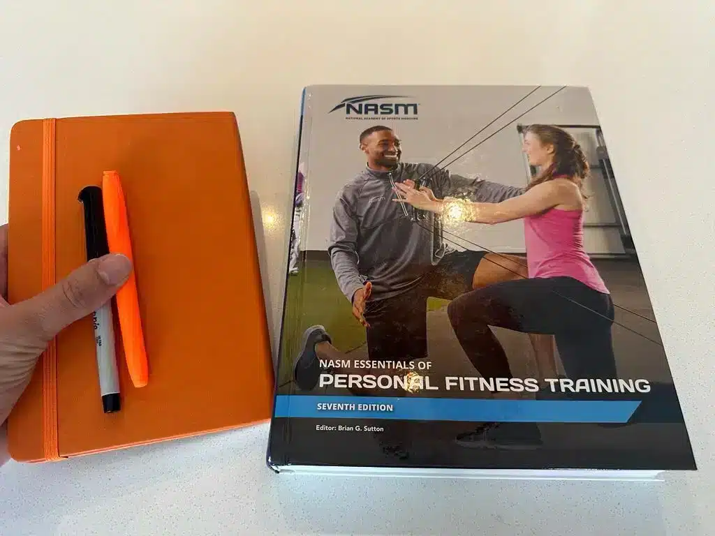 NASM cpt Personal fitness training textbook cover with a journal, sharpie, and highlighter. How much does NASM cost?